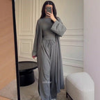 A woman takes a mirror selfie wearing a matching gray two-piece outfit with a long-sleeve top and wide-leg pants, along with a long open-front cardigan. The setting is a modern room with a white paneled wall, a black side table, and a white textured stool.