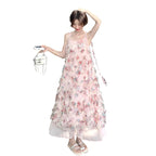 A woman stands outdoors on a paved street in front of a building, wearing a long light pink sleeveless dress covered in 3D floral embellishments. She is holding a small white handbag with a chain strap.