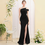 Woman wearing a black one-shoulder dress with a slit, standing in a decorated room with flowers and a chair.