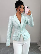 A woman takes a mirror selfie wearing a fitted, light mint green jacquard blazer with white pants. The blazer features a subtle floral pattern, structured shoulders, and a single front button.