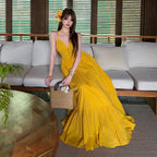 A woman wearing a long, sleeveless yellow maxi dress with a ruffled hem, sitting on a white sofa in a modern living room setting. She is holding a woven straw handbag and has a yellow flower in her hair.