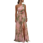 A woman models a one-shoulder pink maxi dress with a green floral print and a large rose detail at the waist, standing on a cobblestone street in a Mediterranean village with white buildings and the blue sea in the background.