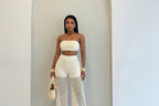 A woman modeling a two-piece matching set consisting of a strapless tube top and high-waisted wide-leg pants with a crochet mesh pattern and fringe hem. She is holding a small woven handbag.
