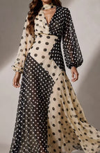 A woman wearing a long-sleeve maxi dress with a black and beige polka dot patchwork design, photographed against a neutral studio background.