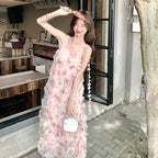 A woman wearing a light pink floral tulle maxi dress with 3D floral appliqués and a long flower-trimmed strap, holding a small white handbag. She is standing outdoors in front of a storefront with a tree trunk on the right.
